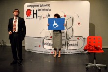 Extrait du spectacle "Embarquez-les !" Une femme tient un panneau avec un pictogramme de personne en fauteuill roulant tandis qu'un homme s'exprime. Tous deux sont devant un paravent "accompagnement du handicap dans l'entreprise".